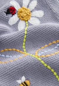 Tissu gris tricoté avec une marguerite blanche brodée, centre jaune, tige verte et motifs de feuilles cousus en orange et bleu, avec une coccinelle rouge sur un pétale.