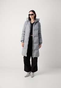 Grijze gewatteerde jas met een capuchon, voorzien van een ritssluiting aan de voorkant en elastische manchetten. Gepaard met zwarte wijde broek en gemusterde hoge hakken.