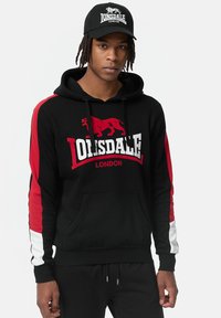Sudadera negra con rayas laterales rojas y blancas, con un gráfico de león y el texto "Lonsdale London". Fabricada en algodón y con un bolsillo frontal.