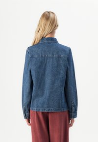 Blonde woman wearing a buttoned denim jacket and loose red pants, seen from the back against a plain white background.