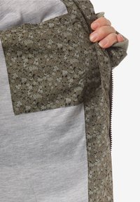 Grön blommönstrad tygöverdrag på ett grått plagg, med en ficka med dragkedja och texturerade detaljer på kanterna.