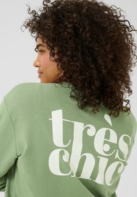 Zelena dukserica od mekog materijala, s velikim bijelim natpisom "très chic" na leđima. Opušteni kroj s vidljivim detaljima šavova.