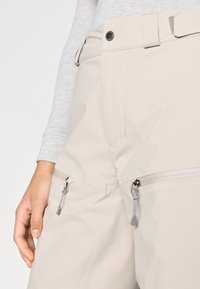 Svetlo bež hlače iz gladkega, vodoodpornega blaga. Imajo dva stranska žepa z zadrgo in kovinsko opremo ter zanko za pas.