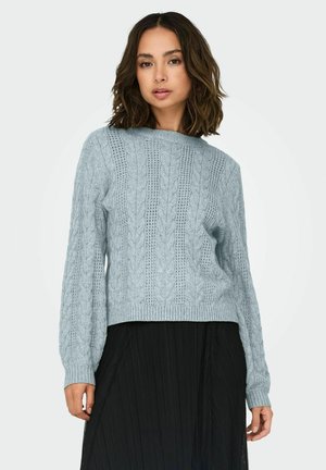 Pull en tricot bleu clair avec un motif en câble, encolure ronde et poignets côtelés, porté sur une jupe plissée noire. Tissu texturé.