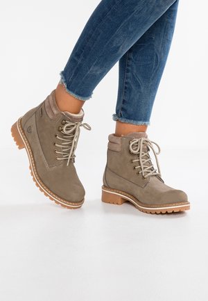 Bruine veterlaarsjes met een doorboorde rand en gevoerde kragen, gedragen bij blauwe jeans met een rafelige zoom, geposeerd tegen een witte achtergrond.