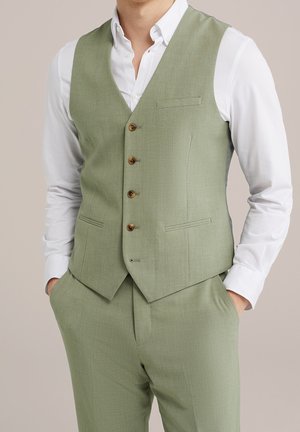 Homme portant un gilet et un pantalon de costume vert clair avec une chemise blanche, les mains dans les poches, debout devant un fond beige uni.