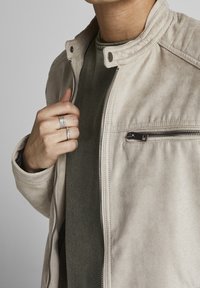 Béžová semišová bunda se stojáčkem, zipovým zapínáním a boční kapsou. V kombinaci se zelenou strukturovanou košili. Na ruce výrazné prsteny.