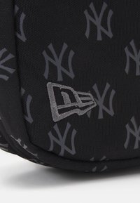 Svart stoffbag med teksturert overflate, grå New York Yankees-logo mønster, og en brodert grå aksentlogo på siden.