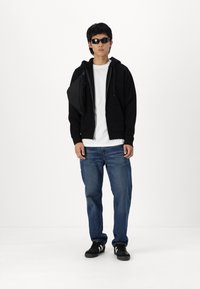 Zwarte rits hoodie, witte t-shirt, blauwe jeans, zwarte sneakers met witte strepen en zwarte zonnebril. Houdt een zwarte tas op de schouder.