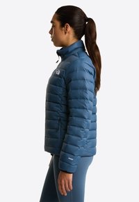 Young woman with dark hair in a ponytail wearing a blue quilted puffer jacket and blue leggings standing in profile.