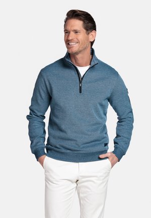 Man glimlachend, draagt een blauwe trui met een kwartritssluiting over een wit shirt en witte broek, handen in de zakken, tegen een effen achtergrond.