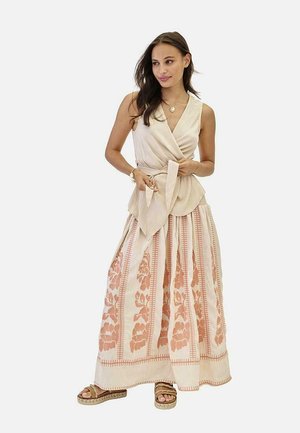 Mujer con blusa beige sin mangas con cierre cruzado y falda larga crema con estampado floral en tonos durazno, llevando sandalias y joyería dorada, parada frente a un fondo blanco.