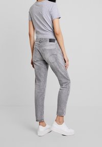 Jeans de mezclilla gris con un corte relajado, textura desgastada, dos bolsillos traseros y un parche negro con el logo en la cinturilla, combinados con zapatillas blancas.