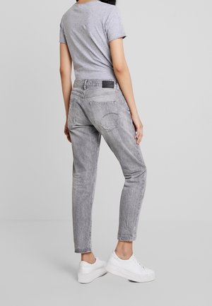 Jeans de mezclilla gris con un corte relajado, textura desgastada, dos bolsillos traseros y un parche negro con el logo en la cinturilla, combinados con zapatillas blancas.