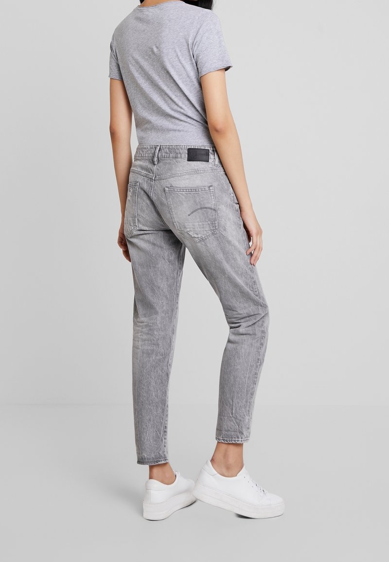 Jeans de mezclilla gris con un corte relajado, textura desgastada, dos bolsillos traseros y un parche negro con el logo en la cinturilla, combinados con zapatillas blancas.