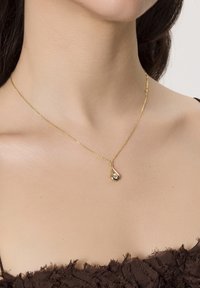 Zbliżenie na szyję kobiety, na której znajduje się delikatny złoty naszyjnik z wisiorkiem w kształcie kropli i małym, błyszczącym kamieniem szlachetnym.