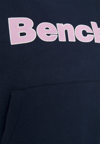 Mørkeblå hoodie med et stort, pink prikket "Bench" logo. Indeholder en frontlomme og er lavet af blødt, struktureret stof.