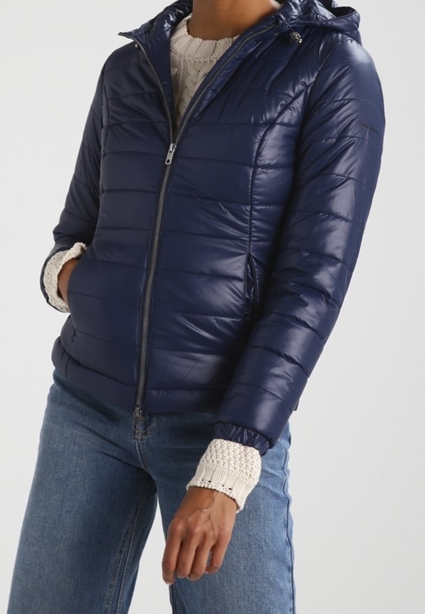 Marineblauer Puffer-Jacke mit Reißverschluss und verstellbare Kapuze. Verfügt über ein gestepptes Muster und elastische Bündchen. Getragen über einem cremigen Strickpullover.