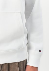 Gros plan sur une personne portant un sweat-shirt blanc avec une poche avant et un petit logo rectangulaire rouge, blanc et bleu sur le poignet.