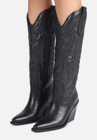 Černé kožené kovbojské boty s propracovaným šitím, špičatou špičkou a vysokým sloupkovým podpatkem. Doplňky zdobí reliéfní vzory.