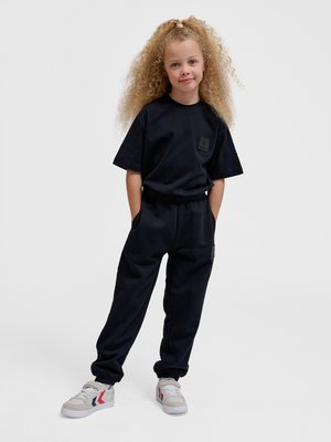 Kind met krullend haar, gekleed in een zwart T-shirt en joggingbroek met witte sneakers, staat met handen in de zakken tegen een effen achtergrond.