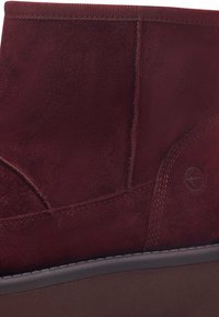 Stivale alla caviglia in suede bordeaux con texture liscia, dettagli cuciti e punta arrotondata. Presenta una suola in gomma marrone e un logo impresso.