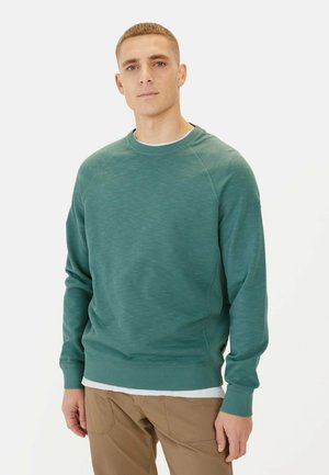 Teal sweatshirt with a textured surface, ribbed cuffs and hem, and a round neckline. Worn over a white t-shirt and paired with khaki pants.