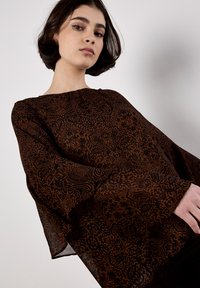 Blusa marrón de estampado con un corte relajado, mangas largas de campana y una tela suave y ligera que presenta intrincados diseños florales.