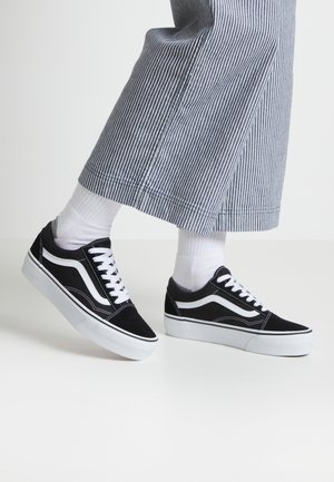 Person iført sorte og hvide sneakers med hvide snørebånd og sokker, kombineret med blå og hvide lodrette stribede bukser med vide ben.
