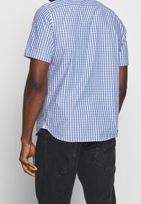 Korte mouwen, blauw-wit geruite shirt met een gebogen zoom, rugpanddetails en zijsplitten, gedragen met zwarte jeans.