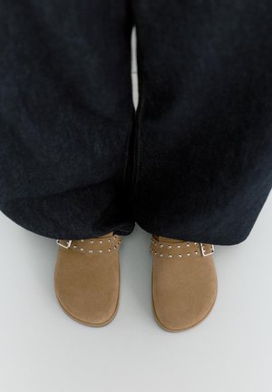 Noge u bež kožnim cipelama bez vezica sa srebrnim zakovicama i crne široke hlače stoje na svijetlosivom podu.