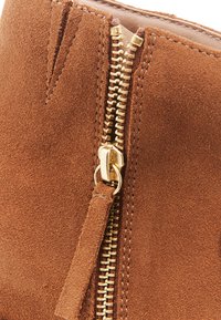 Bottine en daim marron avec une fermeture éclair dorée, des coutures latérales et une finition texturée. Le tirant de la fermeture éclair comprend un détail en tissu avec un gland.