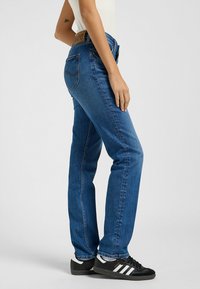 Vaqueros de mezclilla azul con corte recto, que presentan un parche de cuero marrón en la parte trasera de la cinturilla y sutiles detalles de costura. Llevados con zapatillas negras.