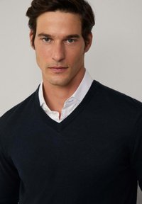 Marineblauer V-Ausschnitt-Pullover aus Strickstoff, über einem weißen Hemd mit Kragen getragen, mit klassischer Passform und glatter Textur.