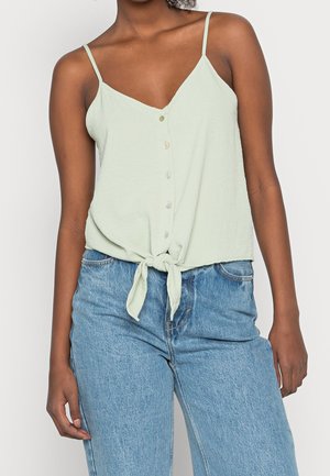 Personne portant un débardeur vert clair boutonné avec un nœud à l'avant et un jean taille haute bleu clair sur un fond uni.