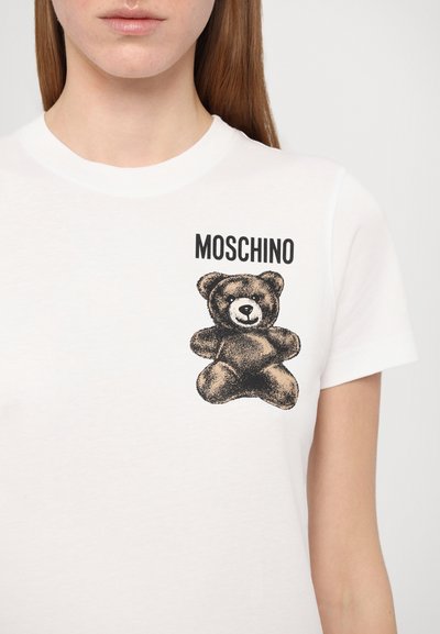Camiseta de algodón blanca con un gráfico de un oso negro y el texto "MOSCHINO". Cuello redondo y mangas cortas con una textura suave.