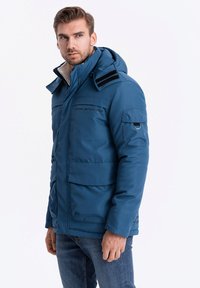 Modrá izolovaná bunda s kapucí, několika kapsami a černými akcenty. Materiál je hladký s prošívanou texturou pro teplo.