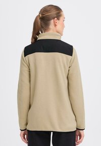 Fleece jack in beige met een zwart schouderpaneel, voorzien van een hoge kraag en contrasterende zwarte zoom. Glad textuur, casual ontwerp.