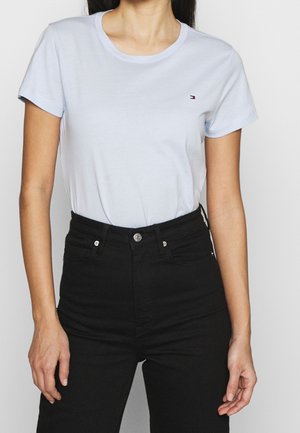 Vrouw draagt een effen wit T-shirt met korte mouwen, ingestopt in een zwarte high waist jeans tegen een effen achtergrond.