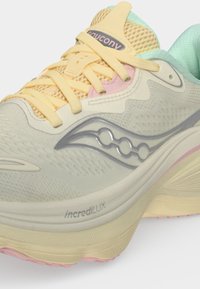 Κοντινή λήψη ενός αθλητικού παπουτσιού Saucony σε παστέλ χρώματα με κίτρινες κορδόνια και τη λέξη "incrediLUX" στη ανοιχτό μπεζ σόλα.