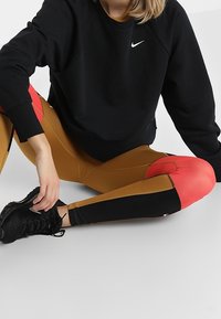 Kobieta siedząca w pozycji skrzyżowanej, ubrana w czarną bluzę Nike, brązowe legginsy z czerwonymi i czarnymi panelami oraz czarne buty sportowe.