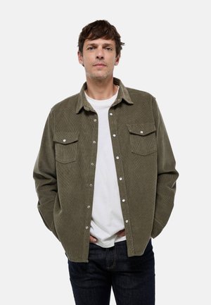 Chemise en velours côtelé vert olive avec un bouton sur le devant, deux poches poitrine et un col évasé, portée sur un t-shirt blanc et un jean bleu.
