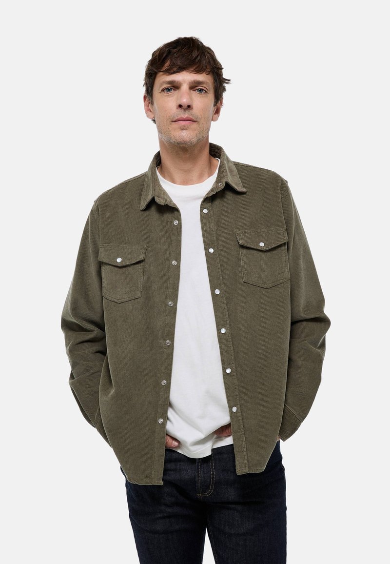 Chemise en velours côtelé vert olive avec un bouton sur le devant, deux poches poitrine et un col évasé, portée sur un t-shirt blanc et un jean bleu.