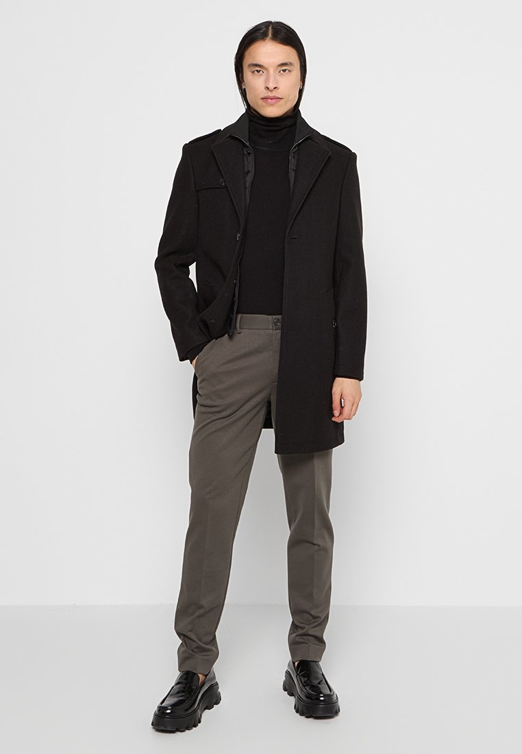 Schwarzer Wollmantel mit maßgeschneiderter Passform, Knopfdetails, kombiniert mit einem schwarzen Rollkragenpullover, grauen Hosen und schwarzen klobigen Schuhen.
