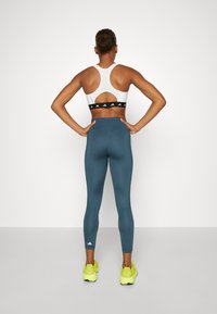 Top sportivo bianco con dettaglio a taglio sulla schiena e fascia nera; leggings blu dal design aderente; scarpe sportive giallo brillante.