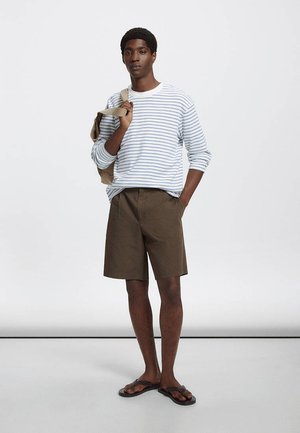 Jeune homme portant une chemise à manches longues rayée, un short marron et des tongs, tenant un sac à dos beige sur une épaule.