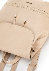 Sac à dos en faux cuir beige avec des accents en clous dorés, une poche avant et des bretelles réglables. Texture lisse avec des détails cousus.