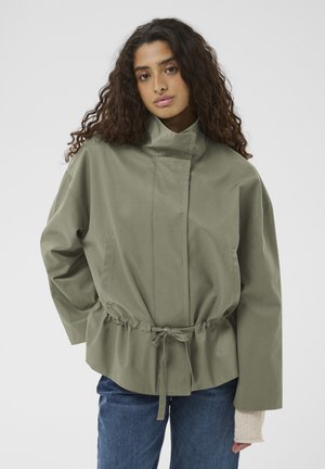 Mladá žena s dlhými kučeravými vlasmi má na sebe nadmernú svetlo olivovú bundu s vysokým golierom a šnúrkou v páse, ktorú dopĺňajú modré džínsy.