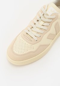 Beige bőr és velúr sportcipők, perforált orral, világos fűzőkkel és textúrált talppal. Egyszerű dizájn és logó részlet.
