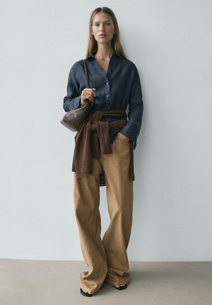 Žena v tmavě modré košili na knoflíky, hnědých širokých kalhotách, hnědém svetru svázaném v pase, držící hnědou kabelku přes rameno, stojící před jednobarevnou zdí.